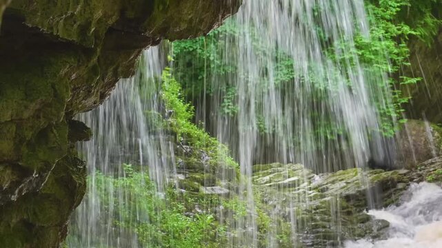 Scenic footage of a beautiful, cascading waterfall flowing over mossy rocks and lush green vegetation in the remote Kara-Syrt Ridge area of Dagestan, within the majestic Caucasus Mountains. This seren