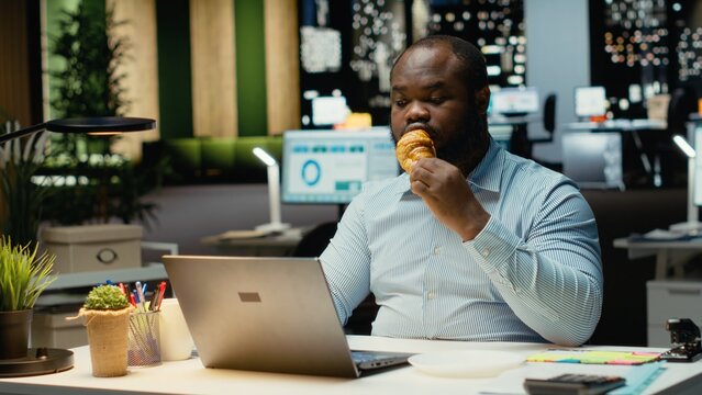 African american male worker struggles with eating disorder in office at night, overeating and being in tension from intense workload. Living unhealthy with junk food and negative emotions. - Powered by Adobe