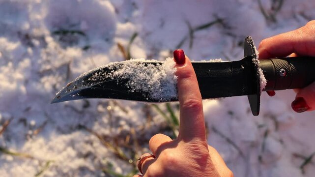 Close-up of a hand holding a knife, its blade covered with freshly fallen snow, highlighting the texture contrast against a snowy winter backdrop. Captures a striking, cold, edgy atmosphere.