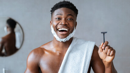 A man stands in a bathroom holding a razor with a joyful expression. He has a towel wrapped around his body and shaving cream on his face. The scene shows a morning grooming routine.