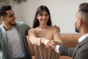 Fototapeta premium Successful Marital Therapy. Happy Millennial Arab Couple Shaking Hands With Family Counselor Thanking For Help And Saved Marriage, Smiling Husband And Wife Sitting On Couch Sofa, Selective Focus