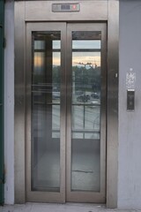 Modern glass elevator with stainless steel doors and city traffic visible through windows. Urban architecture and public infrastructure in Athens, Greece.