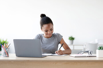 Smiling young african american girl pupil writes in notebook at computer, studies in living room interior, copy space. New normal, schooling education, health care and homework during covid-19 virus