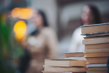A stack of books sits prominently in the foreground while two people are engaged in discussion in the background at a busy place during the day