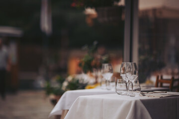 A dining table is set with several glasses placed on a clean tablecloth. The restaurant is located outdoors and shows some blurred background elements in the evening light