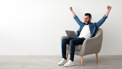 Overjoyed millennial Arab man sitting in armchair with laptop, celebrating online win or work...