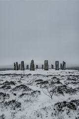 Ancient standing stones stand tall in a snowy field under a vast, overcast sky, evoking a sense of timeless mystery and historical significance.