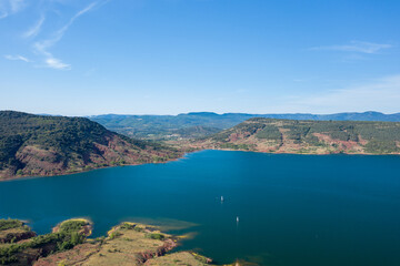 Obraz premium Expansive aerial view of Lac de Salagou with deep blue water bordered by lush green hills and rugged red slopes under a clear sky. The scene is tranquil and open, with distant mountains and a sense of