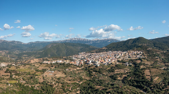 Wide-angle aerial panorama of Orgosolo village nestled among rolling hills, patchwork farmland, and dramatic mountain ridges beneath a bright blue sky in Sardinia.