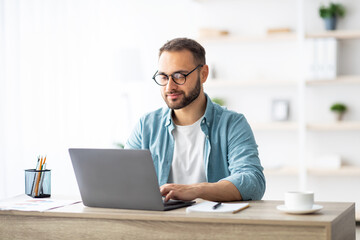Young Caucasian man in eyeglasses working online, sitting at desk and using laptop from home...