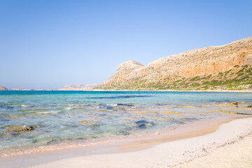 Obraz premium Clear shallow waters meet a sandy shoreline with scattered rocks, backed by sunlit ochre hills and low green vegetation at Balos Beach, Crete. The scene is tranquil, bright, and open under a cloudless