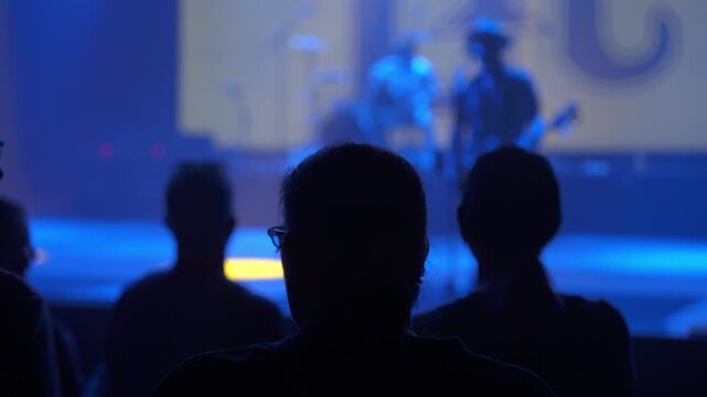 Dark silhouettes of concertgoers fill the foreground while a band performs blurred on stage. Blue lighting and haze create a dramatic live music atmosphere inside the venue.