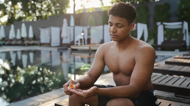 Young mixed race man applying sunblock lotion to his hand, preparing his skin for sun protection near a swimming pool at a resort
