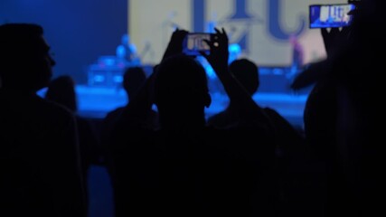 Silhouetted audience members hold up phones to film the stage during a live music performance. Blue lighting and haze surround the crowd while screens capture the show.