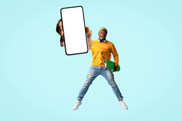 Emotional stylish black guy hipster jumping up with smartphone with blank screen and skateboard on blue, wearing green ice hat and casual outfit, showing nice entertaining app, mockup, copy space
