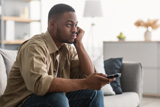 Bored African American Man Watching Boring TV Switching Channels Holding Television Remote Controller Sitting On Sofa At Home. Side View Shot Of Discontented Televiewer
