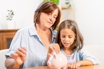 Grandmother teaching granddaughter about saving money while putting coins in piggy bank at home. Generational bonding and family values concept