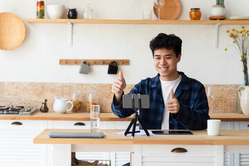 Modern blog, webinar, remote lesson, online chat at home. Happy millennial asian guy gesturing, showing thumbs up, look at smartphone webcam and recording video in modern kitchen interior, free space