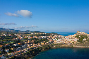 Fototapeta premium Wide aerial view of Castelsardo's modern residential area stretching along the rugged coastline, with pastel buildings, rolling hills, and the historic fortress visible under a clear blue sky.
