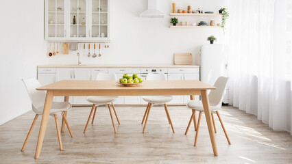 Scandinavian home interior. White and wooden kitchen furniture with various utensils, shelves with plants in pot and dishes, plate with apples on dinning table, panorama