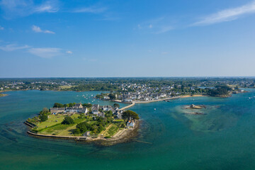 Aerial panorama of a picturesque coastal hamlet on a small island, featuring traditional stone houses, green lawns, and a narrow bridge over turquoise waters in Saint Cado, Brittany. The scene is