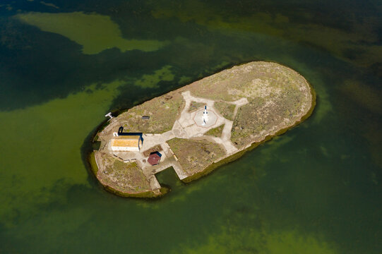 Drone perspective of a small island with a stone chapel and flagpole in the Missolonghi salt lagoon, western Greece. The surrounding green water and geometric paths create a striking, isolated