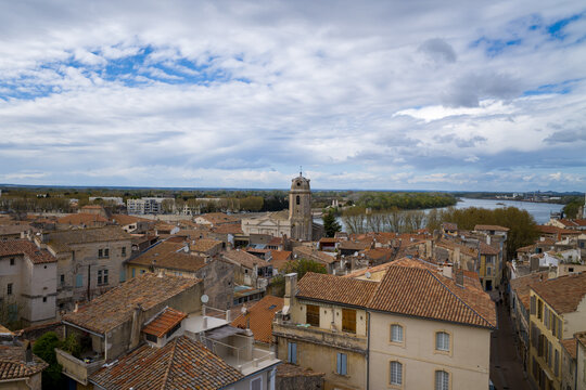 Wide view over the terracotta rooftops of Arles with a historic stone tower and the Rhone River winding through the landscape beneath a dramatic, cloud-filled sky.