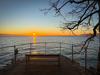 Obraz premium LENS FLARE, SILHOUETTE: Seaside viewpoint with an empty bench overlooks a golden sunset reflected in blue Adriatic Sea. Bare tree branches and a curved metal fence frame the horizon and setting sun.