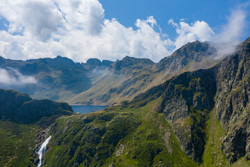 Fototapeta premium A dramatic view of a high-altitude lake nestled among rugged peaks and steep green slopes in the Pyrenees, with a cascading waterfall and drifting clouds under a bright blue sky.