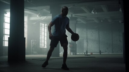 Athletic man dribbling basketball inside dimly lit industrial warehouse during workout