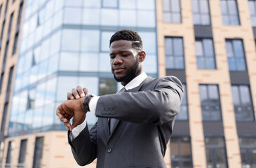 Time management. Focused african american businessman looking at wristwatch, walking during break in downtown area against office building, free space