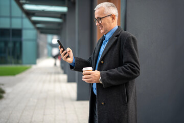 Mature businessman actively using smartphone holding coffee