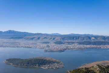Obraz premium Elevated perspective of the tree-covered island of Ioannina set in calm lake waters, with the city of Ioannina and the rugged Pindus mountains forming a dramatic backdrop in Epirus, Greece. Crisp