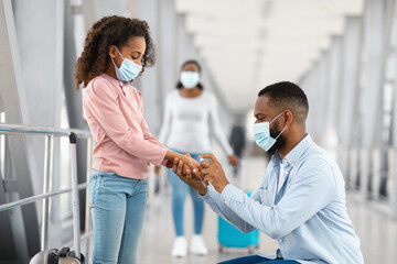 Hygiene Concept. Side view profile portrait of young black man in surgical disposable mask applying antibacterial sanitizer spray on girl's hands, teen cleaning dirty arms in public place at airport © Prostock-studio