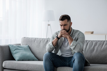 Thoughtful young serious handsome man sit on sofa at home, lost in thoughts, thinking about problem...