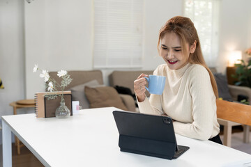 Happy young Asian woman using tablet while enjoying coffee in a cozy living room, remote work and...