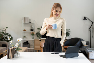 Smiling Asian woman working on digital tablet and holding coffee in modern home office. Concept of...
