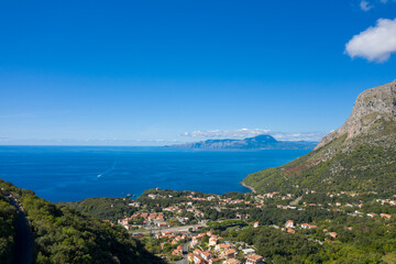 Obraz premium Wide-angle scene showing Maratea village nestled among green hills overlooking the deep blue Tyrrhenian Sea, with rugged mountains in the distance under a clear, sunny sky. The vivid colors and open