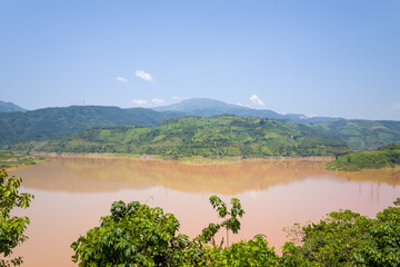 A wide muddy river mirrors lush green hills and terraced farmland beneath a clear blue sky in northern Vietnam. Bright sunlight enhances the vibrant landscape and natural textures.