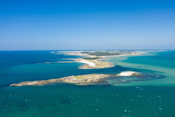 Aerial photograph of the Quiberon peninsula with sandy beaches, rocky outcrops, and a small islet surrounded by vibrant turquoise water under a clear blue sky. The expansive coastline and open sea