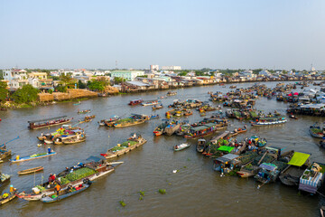Wide daytime view of Cai Rang floating market in Can Tho, Mekong Delta, Vietnam. Dozens of boats loaded with fresh produce and goods gather on the river, surrounded by riverside houses and vibrant