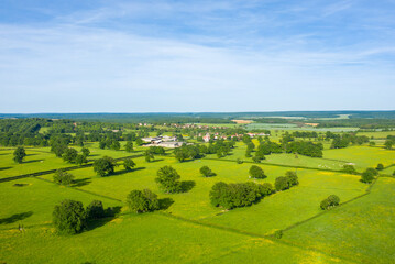 Obraz premium Aerial panorama of lush green fields interspersed with hedgerows and scattered trees, with a small rural village visible in the distance under a clear blue sky near Cuncy les Varzy.
