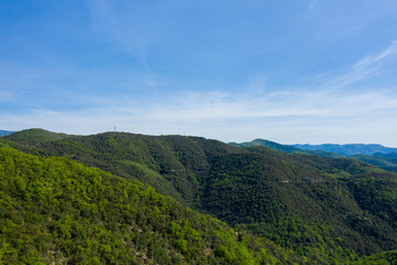 Fototapeta premium Wide aerial view of densely forested hills and valleys stretching beneath a bright blue sky in the Foret domaniale de la Vis. Sunlight enhances the vibrant green foliage and layered mountain landscape