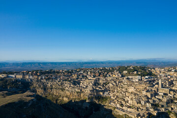 Naklejka premium Expansive aerial view of Matera, Italy, with clustered stone buildings set atop rugged cliffs and rolling hills stretching to the horizon under a vibrant blue sky.