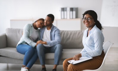 Smiling black female psychologist and young married couple hugging after successful marital therapy at office, free space. Happy psychotherapist after session with reconciled spouses at clinic