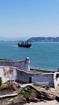 Santo Amaro Da Barra Grande Fortress In Santos Sao Paulo Brazil. Cargo Ship Entering Port With City Skyline In Background. Coast Sky Clouds Seaside Summertime.