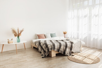 Bedroom interior mockup in boho style with blanket, pillows, white bedding, dried grass and curtain on empty white wall background. Bed, carpet on wooden floor in daylight, table in bedroom interior © Prostock-studio