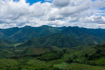 Dense green hills and forested mountain ridges unfold beneath a sky filled with thick, textured clouds in the countryside between Phonsavan and Phou Khoun, Laos. The landscape features undulating