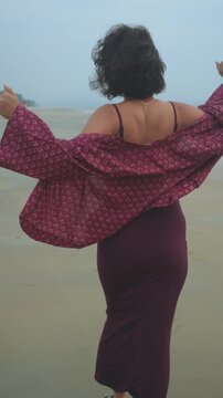 Vertical shot of a woman walking away on a beach, arms raised, birds nearby