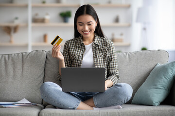 Cheerful young asian woman sitting on couch in living room and using laptop and credit card,...
